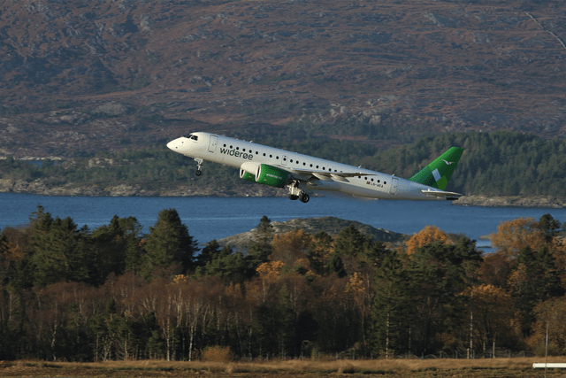 This image shows a Widerøe airplane flying with trees and a lake in the background.